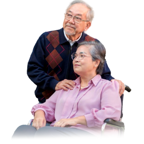 A senior couple — an older man standing behind a woman seated in a wheelchair, both smiling.